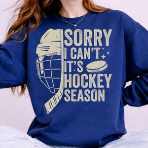 May include: Navy blue sweatshirt with the text "SORRY I CAN'T IT'S HOCKEY SEASON" and a hockey helmet, stick, and puck graphic in beige. The sweatshirt is a comfortable, casual style.