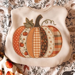 May include: A cream-colored crewneck sweatshirt featuring a large, central pumpkin applique. The pumpkin is constructed from various fabric patches in orange, brown, and cream, showcasing patterns like polka dots, gingham, and floral prints.