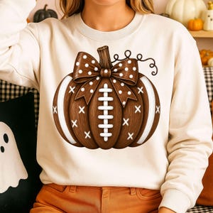 May include: A cream-colored crewneck sweatshirt featuring a large graphic of a brown pumpkin designed to look like a football. The pumpkin has white laces, white stripes on the sides, and a brown bow with white polka dots on the top stem.