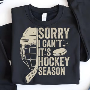 May include: Black sweatshirt with a hockey-themed graphic. The design includes a hockey helmet, stick, and a puck, along with the text "SORRY I CAN'T IT'S HOCKEY SEASON" in a distressed font. The sweatshirt is folded.