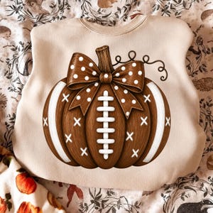 May include: A beige crewneck sweatshirt featuring a large graphic of a brown pumpkin designed to resemble a football. The pumpkin has white laces, white stripes, and a brown bow with white polka dots attached to the stem on the front center.
