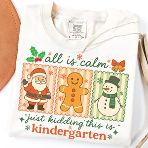 K&ouml;nnte beinhalten: Cremefarbenes T-Shirt mit einem weihnachtlichen Motiv. Das Design zeigt den Weihnachtsmann, einen Lebkuchenmann und einen Schneemann, zusammen mit dem Text "all is calm" und "just kidding this is kindergarten."