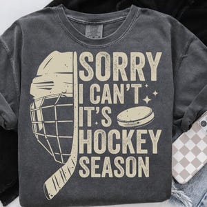 May include: Dark gray sweatshirt with the text "SORRY I CAN'T IT'S HOCKEY SEASON" in a distressed, off-white font. A hockey helmet, stick, and puck graphic are also featured in off-white.