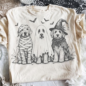 May include: A cream-colored t-shirt featuring a black ink illustration of three dogs dressed for Halloween. One dog is wrapped as a mummy, one is covered by a ghost sheet, and one wears a witch hat and cape. Several small bats fly above.