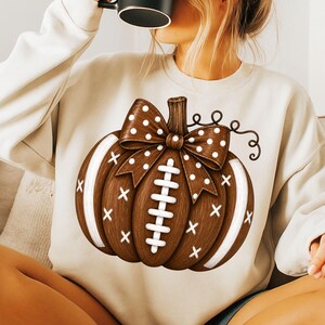 May include: A cream-colored long-sleeve sweatshirt featuring a large graphic of a brown pumpkin designed to resemble a football. The pumpkin has white laces, white stripes on the sides, and a brown bow with white polka dots on the stem.