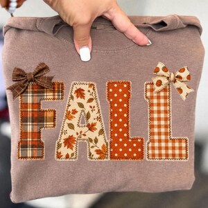 May include: A brown sweatshirt featuring the word FALL appliqued in large letters. Each letter has a distinct pattern: plaid, autumn leaves, white polka dots on orange, and orange gingham with pumpkins. Two fabric bows are attached to the letters.