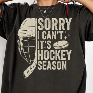 May include: Black t-shirt with a hockey-themed graphic. The design features a hockey helmet, stick, and puck, along with the text "SORRY I CAN'T IT'S HOCKEY SEASON" in a distressed font. A fun and casual top for hockey fans.