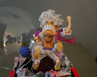 May include: A white-haired anime character figurine with a red and purple cape, sitting on a black and white rock base with a red rim. The character is holding a purple orb in their left hand and has a fierce expression.