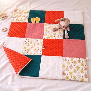 Peut inclure: Une couverture patchwork pour bébé avec une variété de tissus et de couleurs. La couverture présente des carrés de tissus orange, blancs, turquoise et roses. Un jouet lapin en peluche et un anneau de dentition en bois sont sur la couverture.