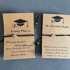 May include: Two graduation-themed bracelets on cards. One card reads "Going Places" with inspirational text. The other says "The Adventure Begins" with a motivational message. Each bracelet has a black cord and a silver charm.