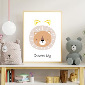 Könnte beinhalten: Ein gerahmter Druck mit einer Cartoon-Löwen-Illustration in Beige und Hellbraun, mit gelben Ohren. Der Text "Dream big" steht darunter. Der Druck befindet sich auf einem weißen Regal mit anderen dekorativen Gegenständen.