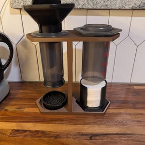 May include: A brown wooden coffee station with two black coffee makers. One has a filter and the other has a lid. The coffee makers are on a wooden surface. White coffee filters are in a black holder.