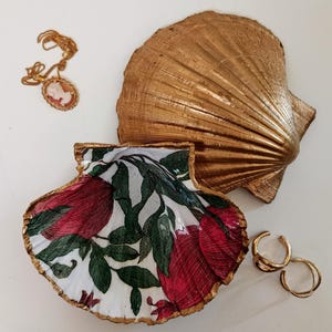 Peut inclure: Un coquillage Saint-Jacques peint en doré avec un intérieur blanc orné d'une illustration de grenade rouge et de feuilles vertes. À côté se trouvent un collier pendentif camée doré et deux anneaux dorés sur fond blanc.