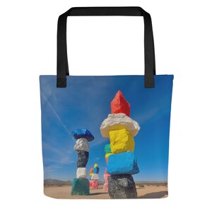 May include: Tote bag featuring a vibrant image of the Seven Magic Mountains art installation in the Nevada desert. The artwork consists of stacked, brightly painted boulders against a clear blue sky. The bag has a black handle.
