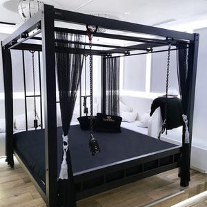 Op de afbeelding: Een zwart metalen bedframe met een hemelbed en zwarte, doorschijnende gordijnen. Het bed heeft een zwarte matras en zwarte kussens met gouden letters die "LOXXY" lezen. Er hangen kettingen aan de hemel.