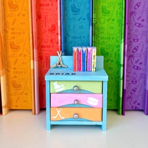 May include: A small, light blue wooden nightstand with three drawers, each painted a different color: green, pink, and orange. The drawers feature hockey-themed illustrations. The word "BRIAR" is painted on the front. Books and a hockey stick figurine sit on top.