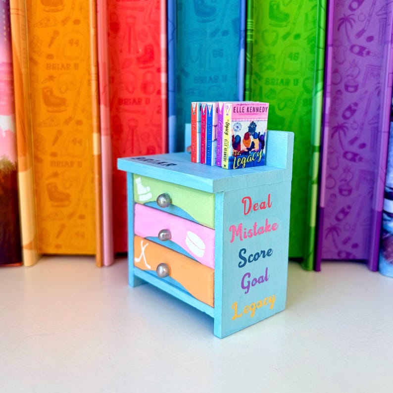 May include: A miniature blue dresser with three drawers, each painted a different color and adorned with silver knobs. The drawers are green, pink, and orange. The text "Deal, Mistake, Score, Goal, Legacy" is written on the side. A stack of books sits on top.
