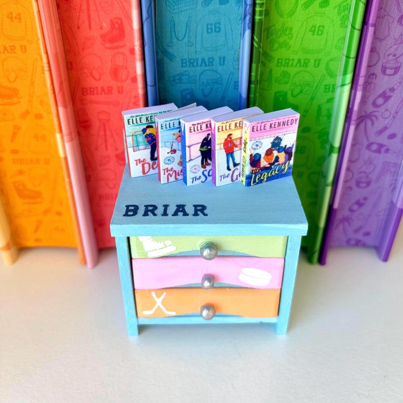 May include: A miniature blue dresser with three drawers, each painted a different pastel color. The top of the dresser has the word "BRIAR" and a row of miniature books. Behind the dresser are colorful books with the text "BRIAR U".