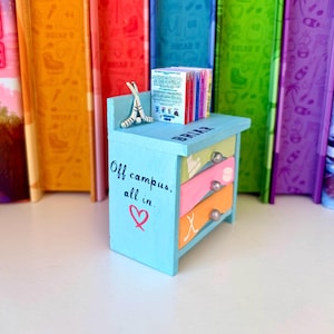 May include: A miniature blue painted dresser with three drawers, each in a different color. The top of the dresser has the word "BRIAR" written on it. The dresser has the text "Off campus, all in." and a heart symbol. A stack of books and a hockey stick figurine sit on top.