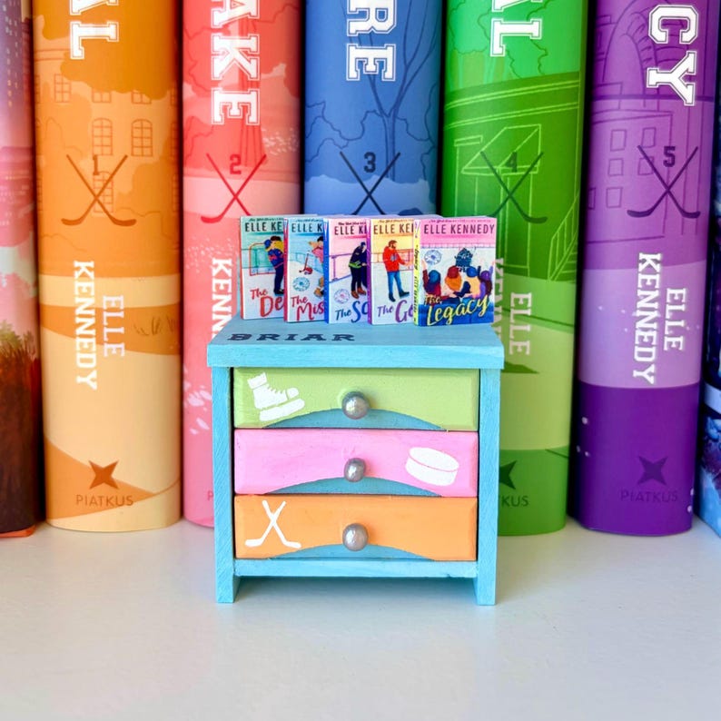 May include: A miniature blue wooden dresser with three drawers, each painted a different color: green, pink, and orange. Each drawer features a hockey-themed design. Small books are displayed on top of the dresser, with larger books in the background.