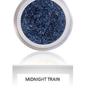 May include: A small, clear jar filled with dark blue, shimmering eyeshadow. The eyeshadow is labeled "Midnight Train" in black text on a white rectangular label below the jar. The jar is set against a white background.