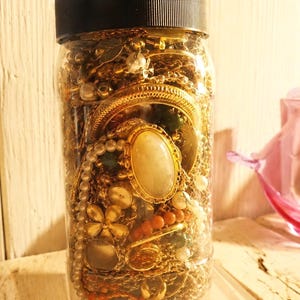 May include: A clear plastic jar with a black screw-top lid filled with a collection of assorted gold-toned jewelry, including chains, a large oval cabochon pendant, pearl-like beads, floral-shaped charms, and small orange coral-colored beads.