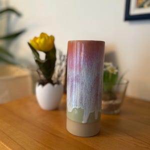 Könnte beinhalten: Eine zylindrische Keramikvase mit Farbverlauf. Die Oberseite ist rot-rosa, geht in Weiß über, dann in Hellgrün und schließlich in einen beigefarbenen Sockel. Die Vase steht auf einer Holzoberfläche.