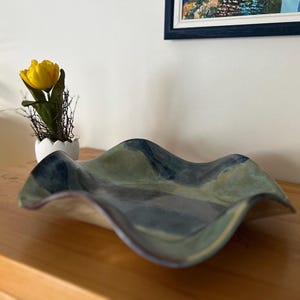Könnte beinhalten: Dekorative Keramikschale mit wellenförmigem Rand und blau-grüner Farbgebung. Die Schale steht auf einer Holzoberfläche, im Hintergrund eine gelbe Blume in einem weißen Topf. Das Design der Schale erinnert an Meereswellen.