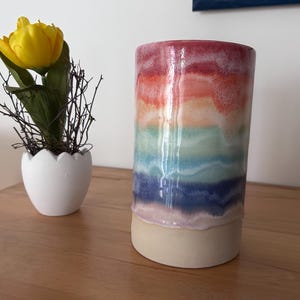 Könnte beinhalten: Eine zylindrische Keramikvase mit Regenbogenverlauf, mit Bändern in Rot, Orange, Grün, Blau und Lila. Die Vase steht auf einem beigefarbenen Sockel. Links steht ein kleiner weißer, eiförmiger Pflanztopf mit einer gelben Tulpe.