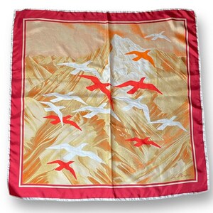 May include: Square silk scarf with a mountain and bird design. The scarf features a red border and a white trim. The background is a mix of gold, orange, and white, with red and white birds flying across the scene.