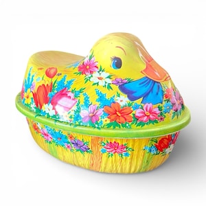May include: A yellow duck-shaped container with a lid, decorated with colorful flowers and a green rim. The duck has a blue bow and an orange beak. The container is likely made of metal and has a vintage or retro aesthetic.
