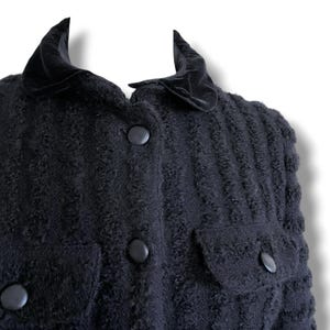 May include: A close-up of a black textured jacket with a velvet collar and large black buttons. The jacket has two pockets and a textured pattern throughout the fabric. The collar is a darker shade of black.