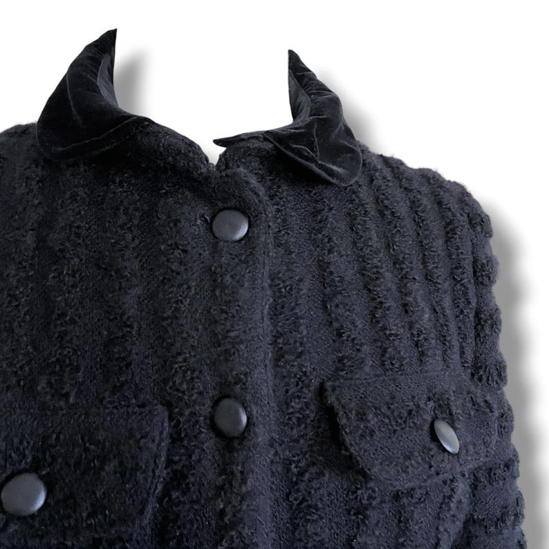 May include: A close-up of a black textured jacket with a velvet collar and large black buttons. The jacket has two pockets and a textured pattern throughout the fabric. The collar is a darker shade of black.