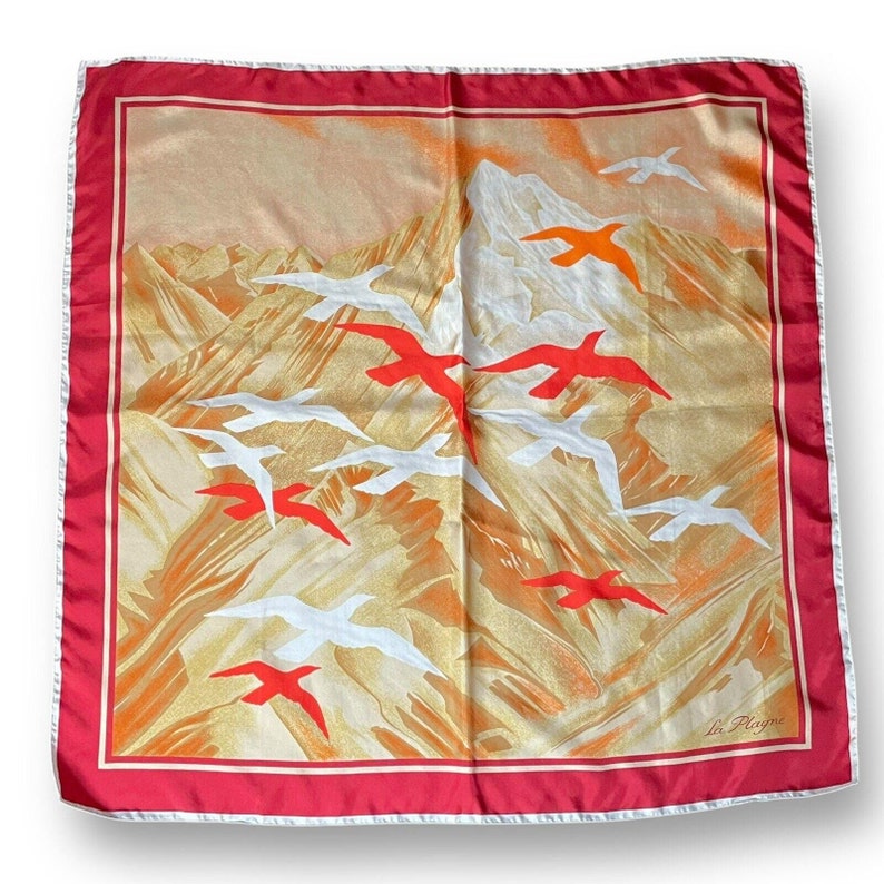 May include: Square silk scarf with a mountain and bird design. The scarf features a red border and a white trim. The background is a mix of gold, orange, and white, with red and white birds flying across the scene.