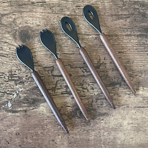 May include: Four black and brown wooden spoons with a unique design. The spoons have a rounded, oval-shaped head with a small hole in the center. The handles are made of dark brown wood.