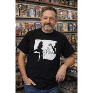May include: A black t-shirt featuring a white graphic of a cartoon character seated at a table. The person is standing in front of a wall of comic books.