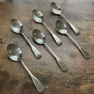 May include: Six stainless steel spoons on a wooden surface. The spoons have oval bowls and long, slender handles. The spoons are reflective, and the wooden surface has a dark, warm tone.