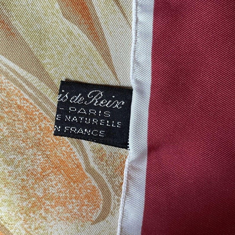 May include: A silk scarf with a label that reads "is de Reix - PARIS - SOIE NATURELLE - FRANCE." The scarf features a mix of colors, including orange, yellow, and red, with a white and red striped border.
