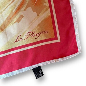 May include: A silk scarf with a vibrant design featuring shades of orange, yellow, and red. The scarf has a red border with a white trim. The words "La Plagne" are written in cursive. A black tag with white text is attached.