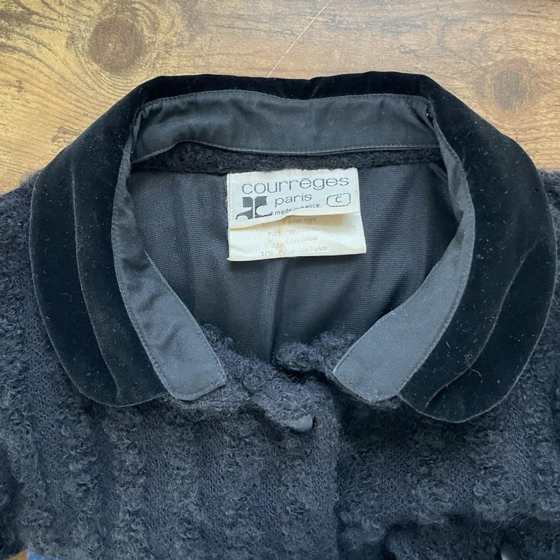 May include: Close-up of a black jacket with a velvet collar. The jacket has a textured, boucl&eacute; fabric. A white tag inside the collar reads "courreges paris made in france". The jacket is a vintage piece.