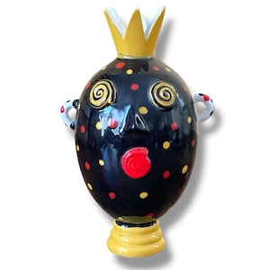 Może przedstawiać: Czarny ceramiczny wazon z fantazyjną twarzą, żółtą koroną i czerwonymi i żółtymi kropkami. Wazon ma okrągły korpus i małą podstawę.
