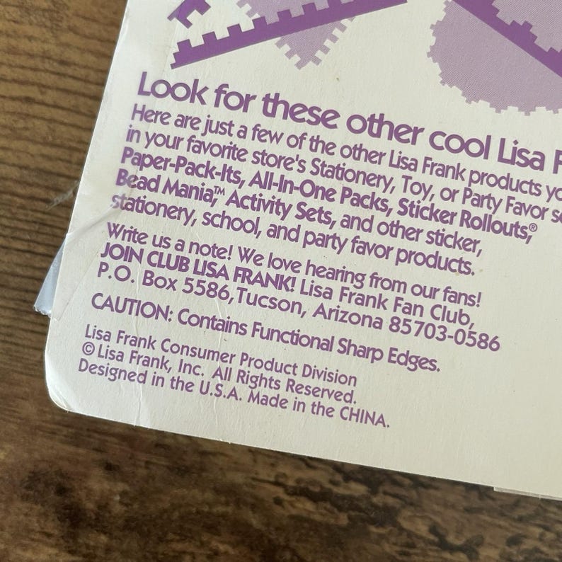 May include: A white product label with purple text promoting Lisa Frank products. The label lists stationery, toys, and party favours. It also includes a caution about functional sharp edges. Made in China.