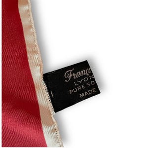 May include: A close-up of a red silk scarf with a cream-colored border. A black tag with the brand name "Franco Lyon" and "Pure Soie Made" is attached to the scarf. The scarf has a smooth, shiny texture.