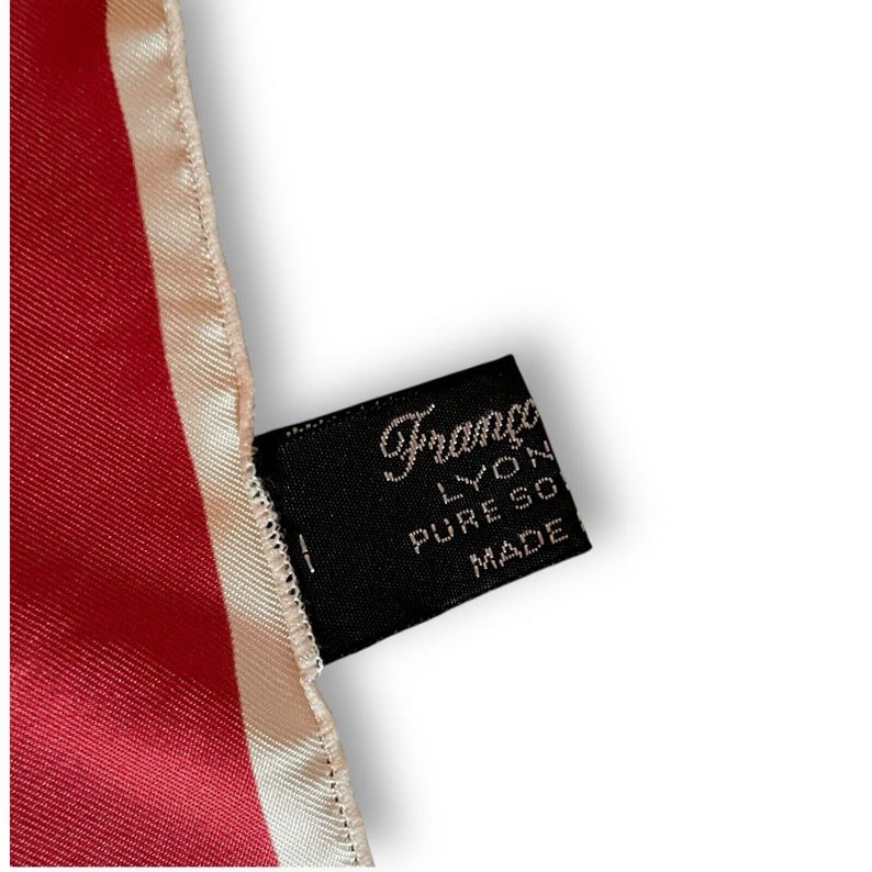 May include: A close-up of a red silk scarf with a cream-colored border. A black tag with the brand name "Franco Lyon" and "Pure Soie Made" is attached to the scarf. The scarf has a smooth, shiny texture.