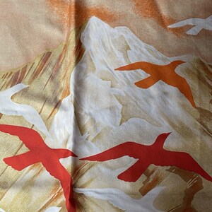 May include: A fabric with a mountain and bird design. The background is a blend of tan, orange, and white, with stylized mountains and red and orange birds in flight. The fabric has a soft, flowing appearance.