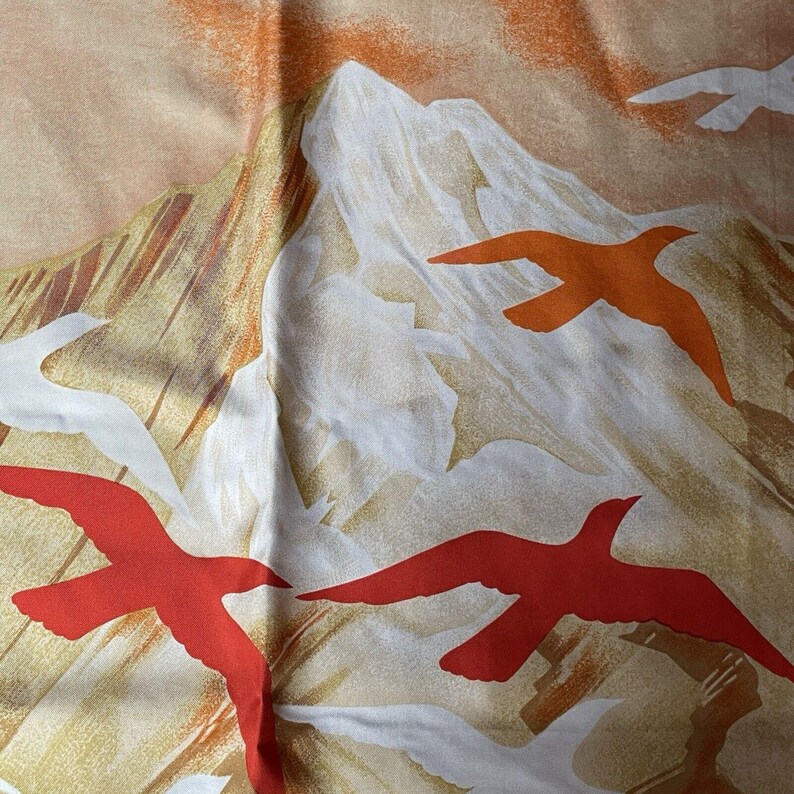 May include: A fabric with a mountain and bird design. The background is a blend of tan, orange, and white, with stylized mountains and red and orange birds in flight. The fabric has a soft, flowing appearance.