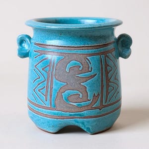 May include: A turquoise ceramic pot with two small handles and a wide rim. The pot features a brown geometric design and a central gray abstract shape. The base has three small feet.
