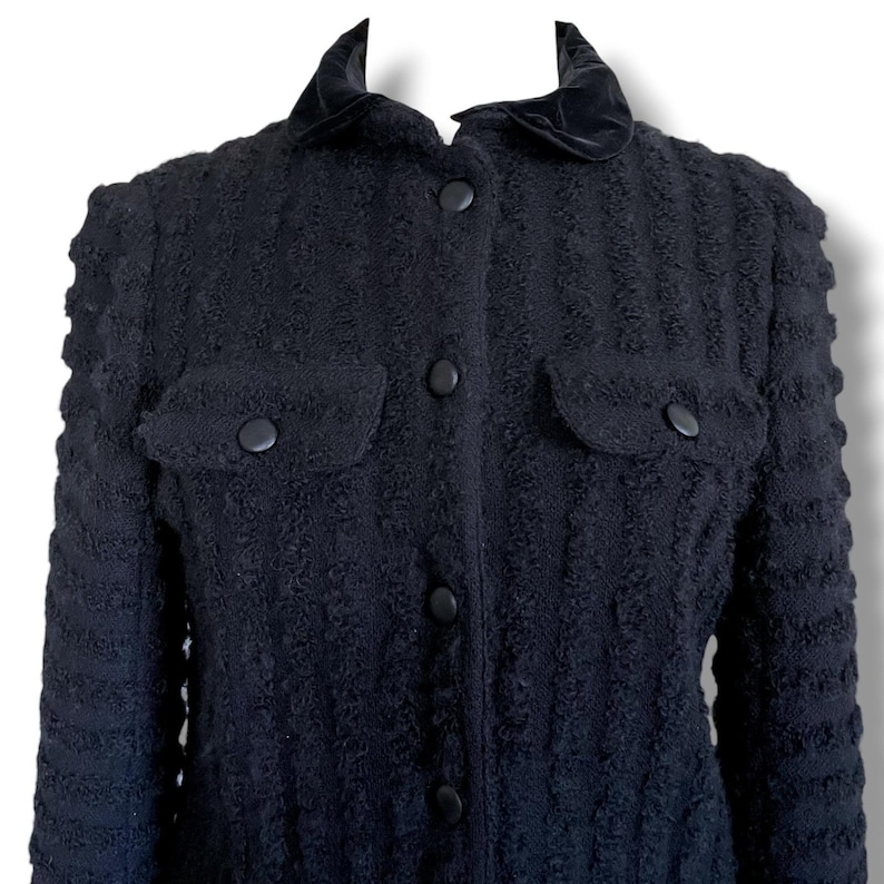 May include: A black textured jacket with a velvet collar and two front pockets. The jacket has a button-up closure and a textured pattern throughout the fabric. The jacket is a classic style.