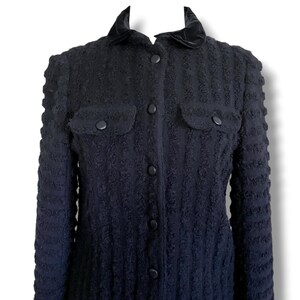 May include: A dark navy blue jacket with a textured fabric. The jacket features a velvet collar, two front pockets, and black buttons down the front. The sleeves have a textured design. A classic outerwear piece.