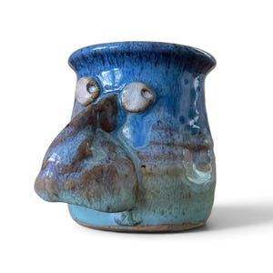 May include: A ceramic mug with a glossy blue glaze featuring a prominent, sculpted brown nose and two small, round, off-white eyes protruding from the front. The mug has a flared rim and a gradient color transition from dark blue to light green.